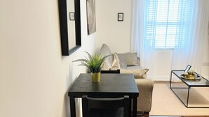 A living room with a sofa, a coffee table, and a dining table with a chair. There is a plant on the dining table and a window with curtains. - Comfortable One Bedroom Apartment London (London)