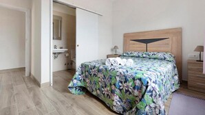 Un dormitorio con cama, cabecera de madera, mesita de noche, lámpara y baño visible a través de una puerta abierta.