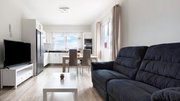 Una sala de estar moderna con un sofá gris oscuro, un televisor de pantalla plana sobre un soporte blanco y una pequeña mesa blanca con una planta.