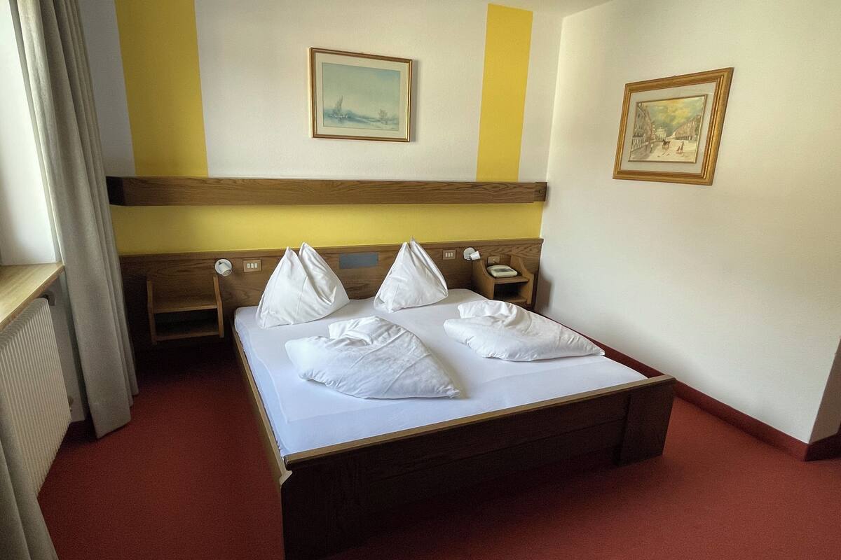 A hotel room with a bed, a bedside table, a nightstand, a window, and two framed pictures on the wall.