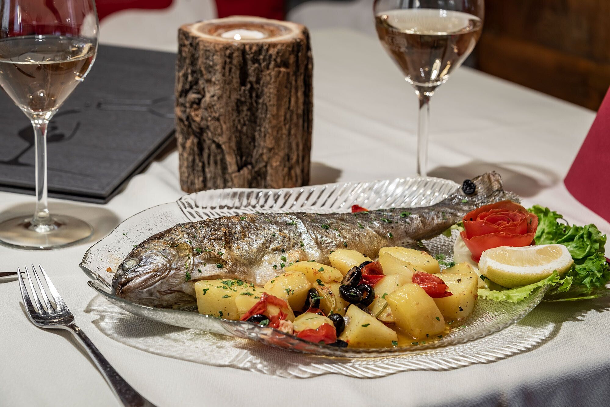 Une assiette de poisson accompagnée de pommes de terre et de légumes, un verre de vin blanc et une bûche de bois.