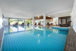 A large indoor swimming pool with clear blue tiles, surrounded by lounge chairs and a view of greenery outside. - Hotel Wiedenhofer (Terento)