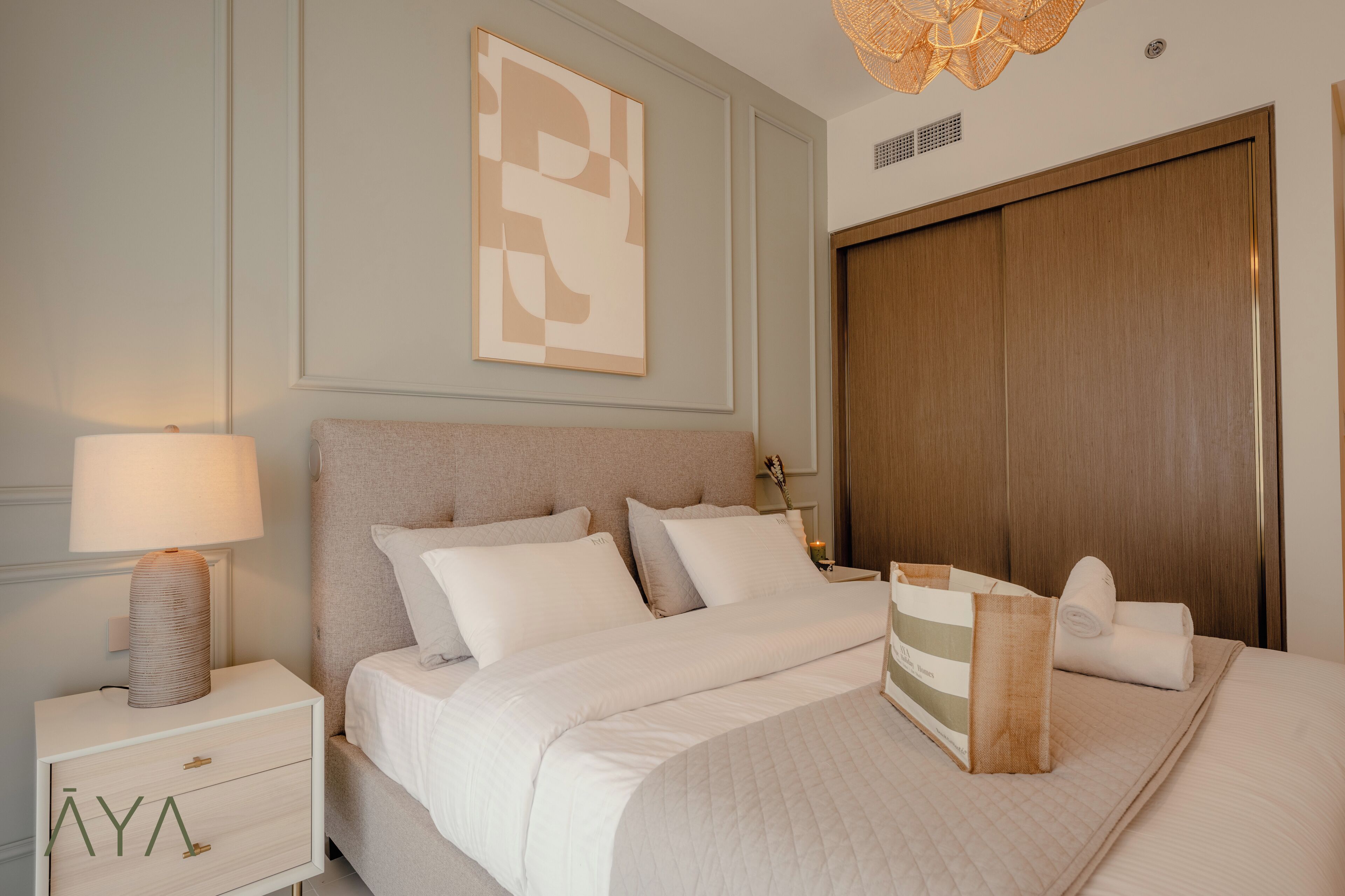 A neatly arranged bedroom with a bed, a bedside table with a lamp, a wooden wardrobe, and a wall-mounted abstract artwork.