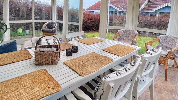 Une véranda avec une table blanche dressée pour quatre personnes, entourée de chaises en osier, et de grandes fenêtres offrant une vue sur le jardin et les maisons voisines.