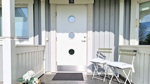 Une porte blanche avec une plaque portant un numéro, une petite fenêtre ronde et un paillasson à l’entrée. Sur le porche, on trouve deux chaises pliantes blanches et un panier en osier rempli de fleurs.