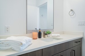 Un baño moderno con un mostrador blanco, un lavamanos, un espejo, un montón de toallas, un dispensador de jabón y una pequeña planta en maceta.