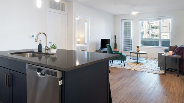 A modern kitchen with a built-in dishwasher, a sink, and pendant lights. There is a dining area with a table and chairs, and a large window providing natural light. - Westerly (Dallas)