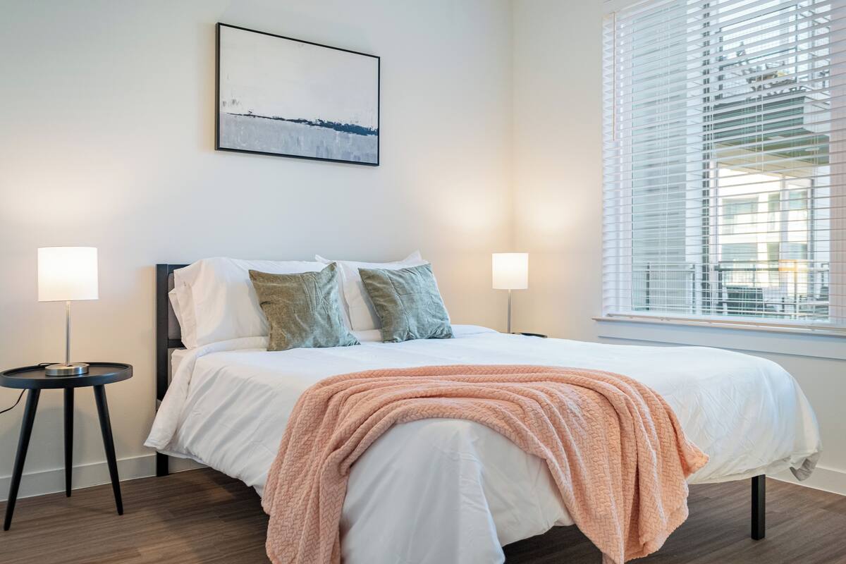 A bedroom with a bed, two bedside lamps, a window with blinds, and a wall-mounted abstract painting.