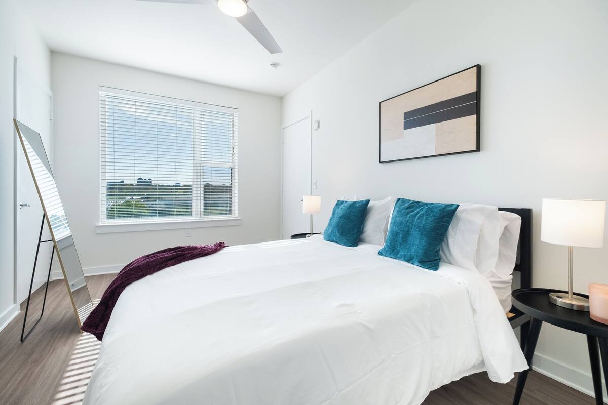 A modern hotel room with a large bed, a standing desk, a window with blinds, and a ceiling fan.