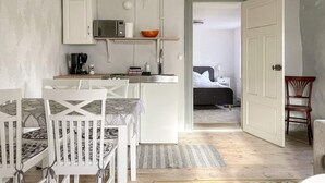 A compact kitchen with white cabinets, a microwave, and a small countertop. There is a dining area with a white table and chairs. A bed is visible in the adjacent room. - 5 Person Holiday Home in Stenkyrka (Tingstade)