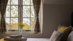 Un dormitorio con cama, ventana con cortinas, un jarrón con flores y una mesita de noche.
