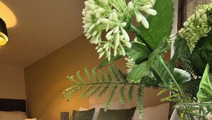 A hotel room with a bed, pillows, a bedside table with a vase of flowers, and a nightstand with a lamp.