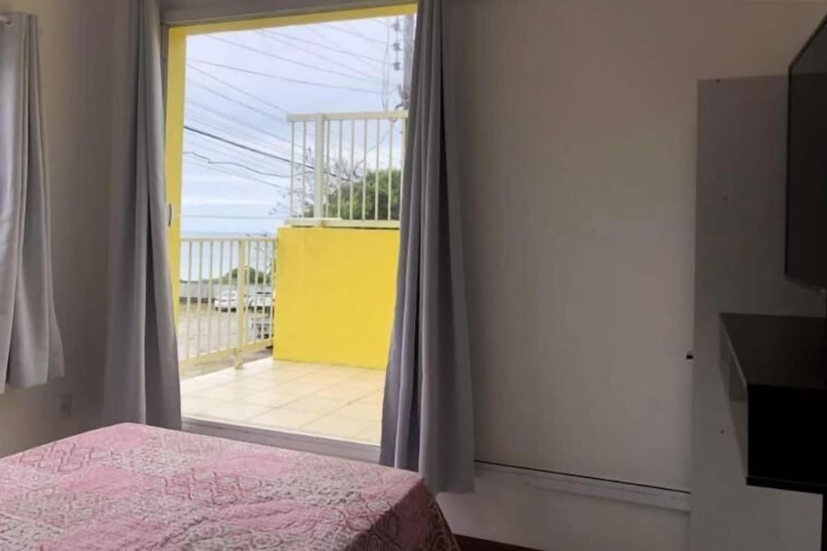 A room with a bed, a TV, a window with curtains, and a view of a yellow building outside.