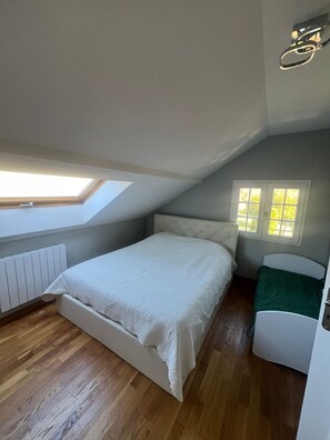 Une chambre avec un plafond en pente, un puits de lumière, un lit recouvert d’une couette blanche, une table de chevet et une fenêtre avec des volets blancs.