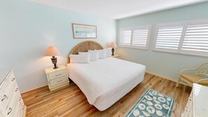 Un dormitorio con una cama grande, dos mesitas de noche con lámparas, un cabecero de mimbre, un cuadro en la pared y una ventana con persianas.