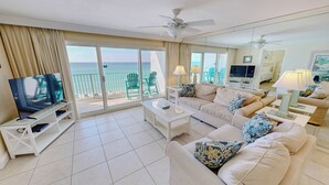 A spacious living room with a TV, white furniture, a balcony with chairs, and a view of the ocean. - Beach House 602A Gulf Front 2 BR Stunning Views and Seasonal Beach Service (Miramar Beach)