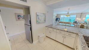 A kitchen with a refrigerator, a trash bin, and a dining area with a view. - Beach House 602A Gulf Front 2 BR Stunning Views and Seasonal Beach Service (Miramar Beach)