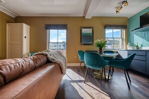 A living room with a brown leather sofa, teal chairs, a dining table, and a kitchen area with a TV.