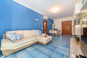 A living room with a beige sectional sofa, a dining area with a table and chairs, and a kitchen area with a wooden cabinet. - Appartamento Sally in Sorrento (Sorrento)