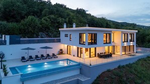 A modern two-story house with a swimming pool, outdoor seating, and a view of a forested hillside. - Luxurious Villa Gabi with an infinity pool and sauna (Mihotici)