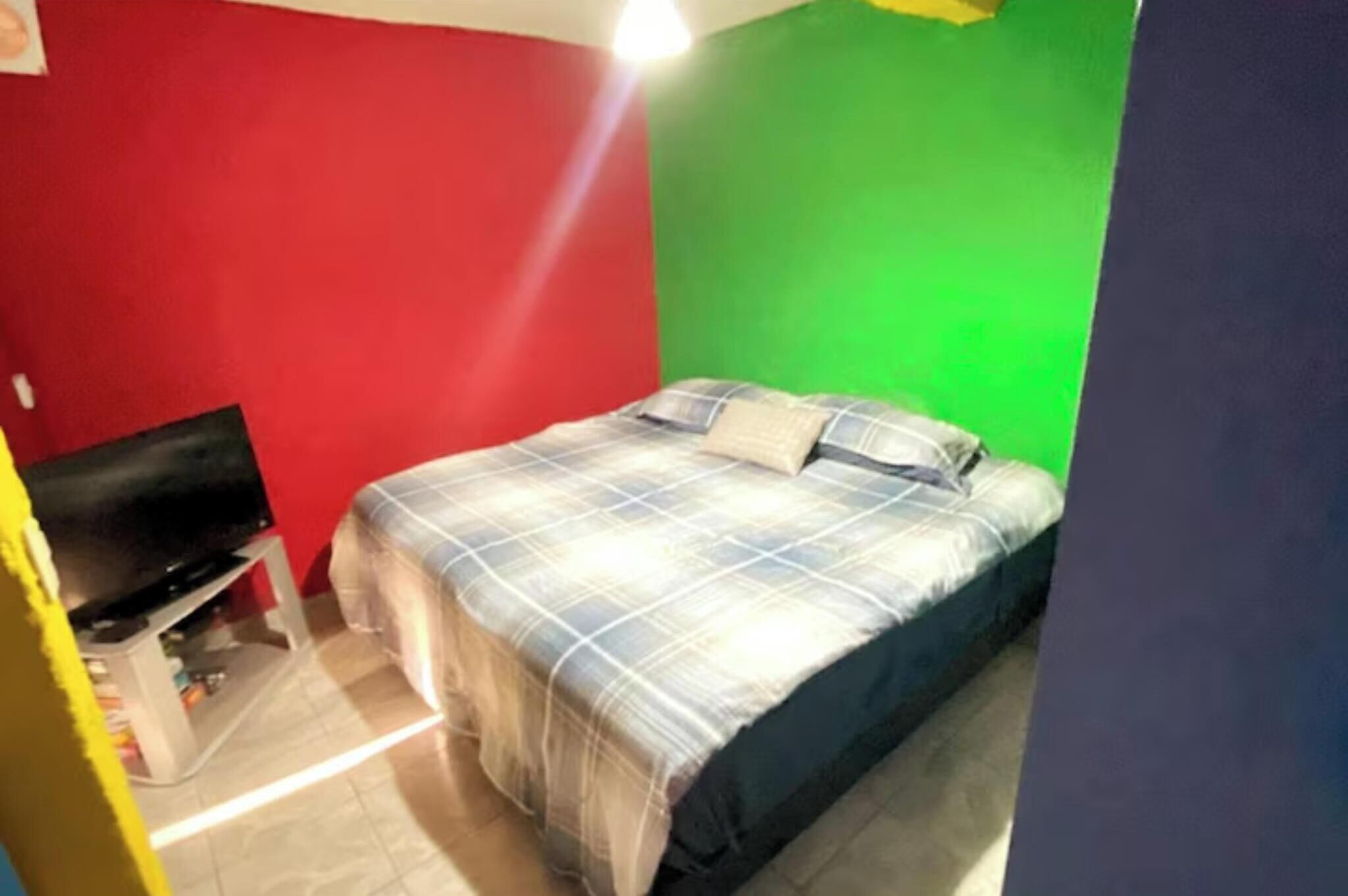 Une petite chambre avec un lit simple, un mur rouge et vert, un téléviseur posé sur un meuble et un lit recouvert d’une couverture à carreaux.