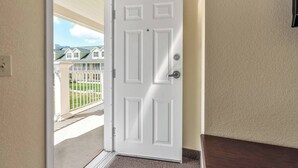 A white door with a doorknob and a mail slot, leading to a covered outdoor area with a view of houses and a lawn.