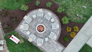Un espace extérieur circulaire aménagé avec des sièges, comprenant un foyer au centre, entouré de chaises et de bancs blancs, ainsi qu'un chemin pavé et des parterres de fleurs jaunes.