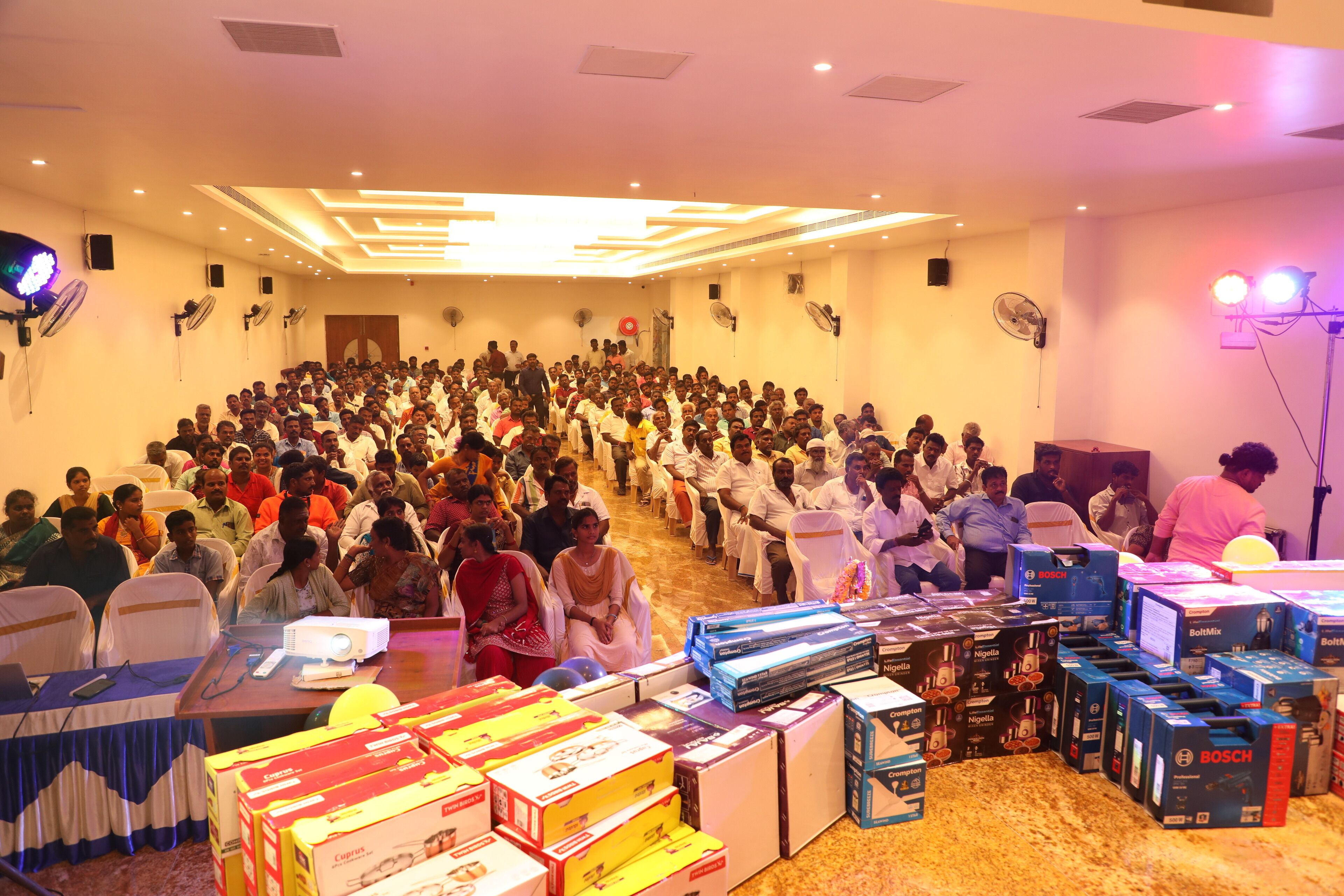 Un salón de conferencias con un público numeroso sentado en filas, un escenario con un atril y diverso equipo electrónico y cajas sobre las mesas.