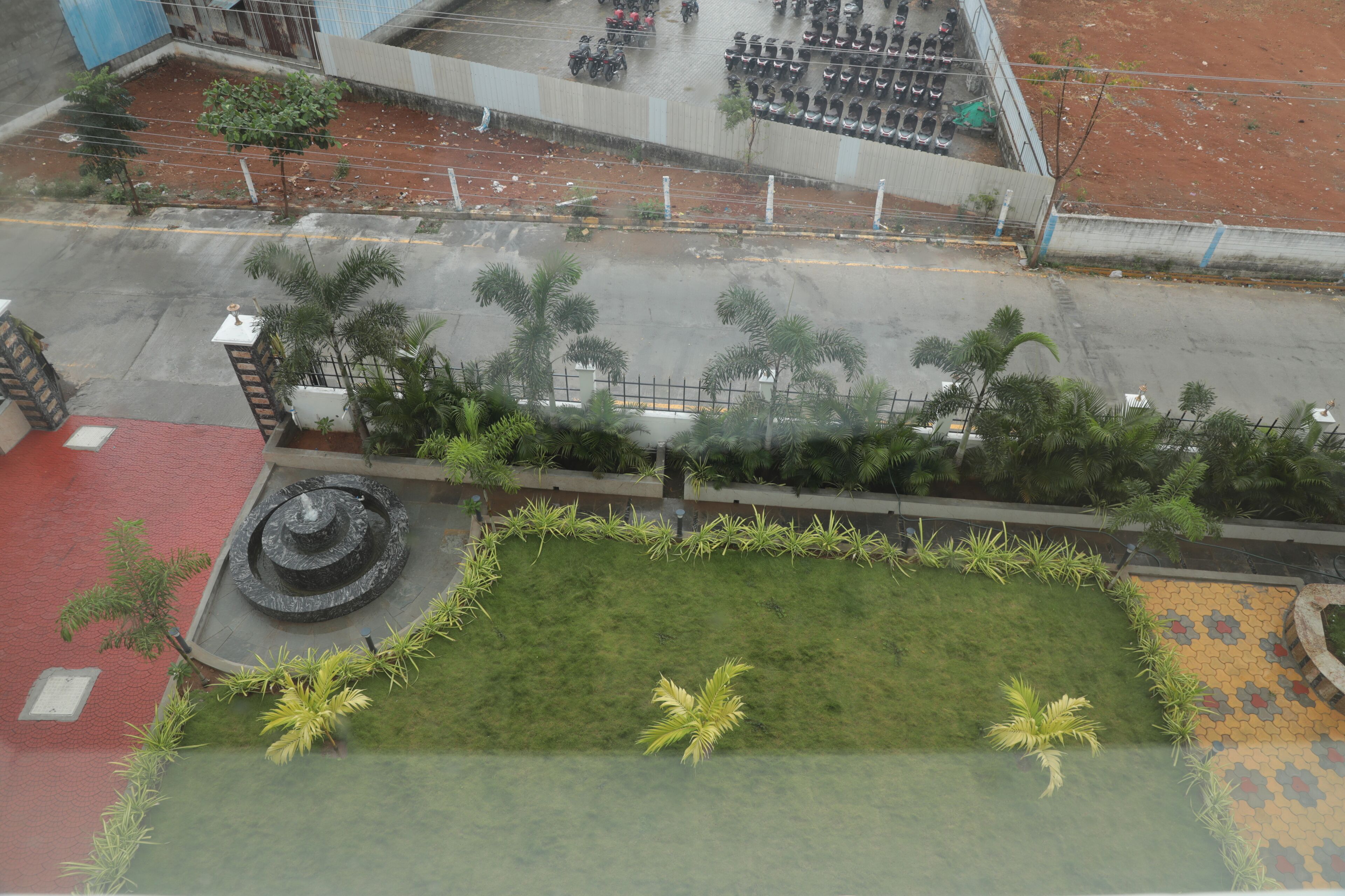 An aerial view of a parking lot with multiple rows of parked vehicles, a grassy area with a circular fountain, and a red building in the foreground.