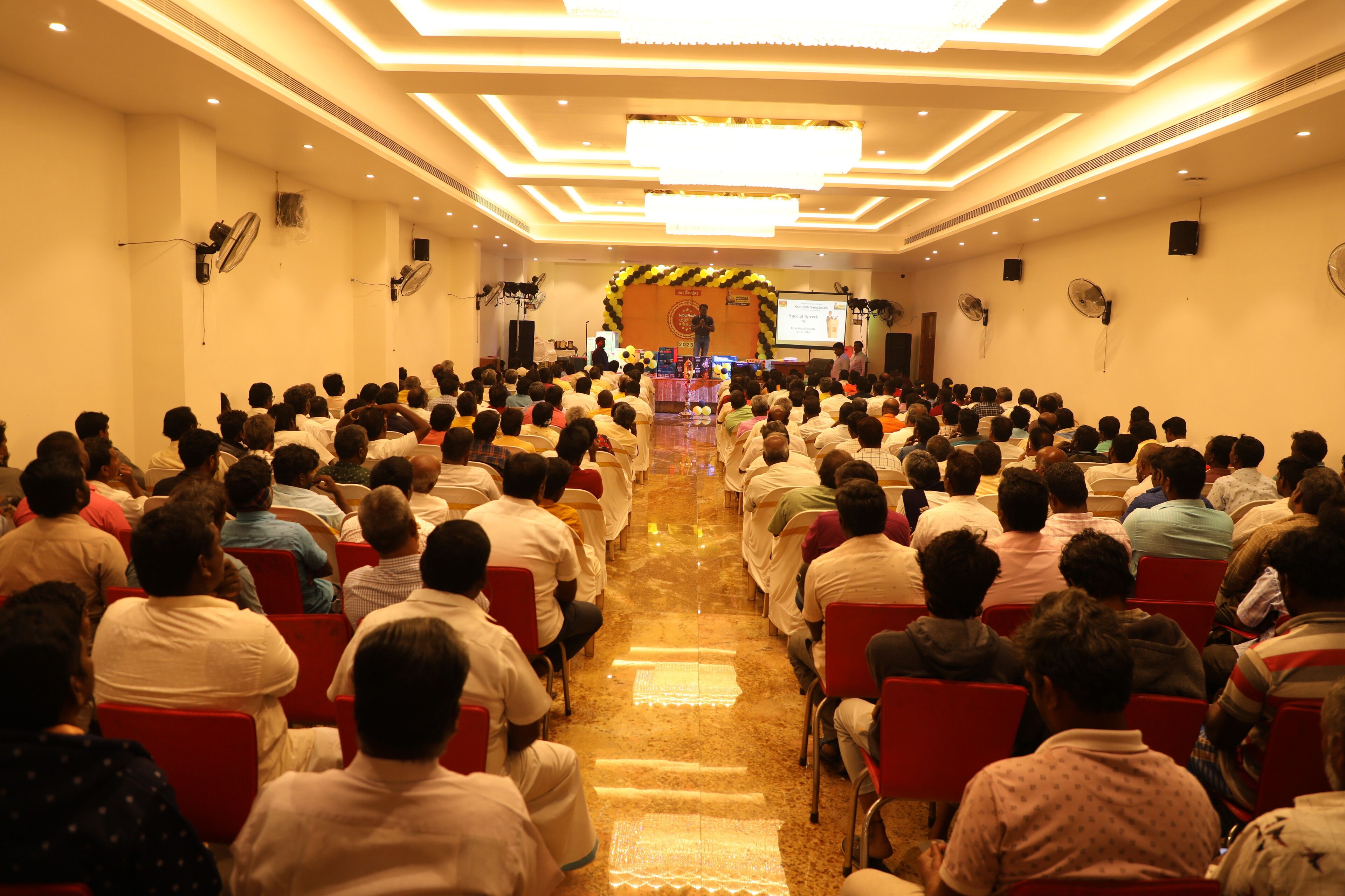 A conference room with rows of chairs facing a stage with a speaker and a presentation screen.