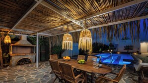 Un patio al aire libre con piso de piedra, mesa y sillas de madera, luces colgantes, chimenea de ladrillo y una piscina.