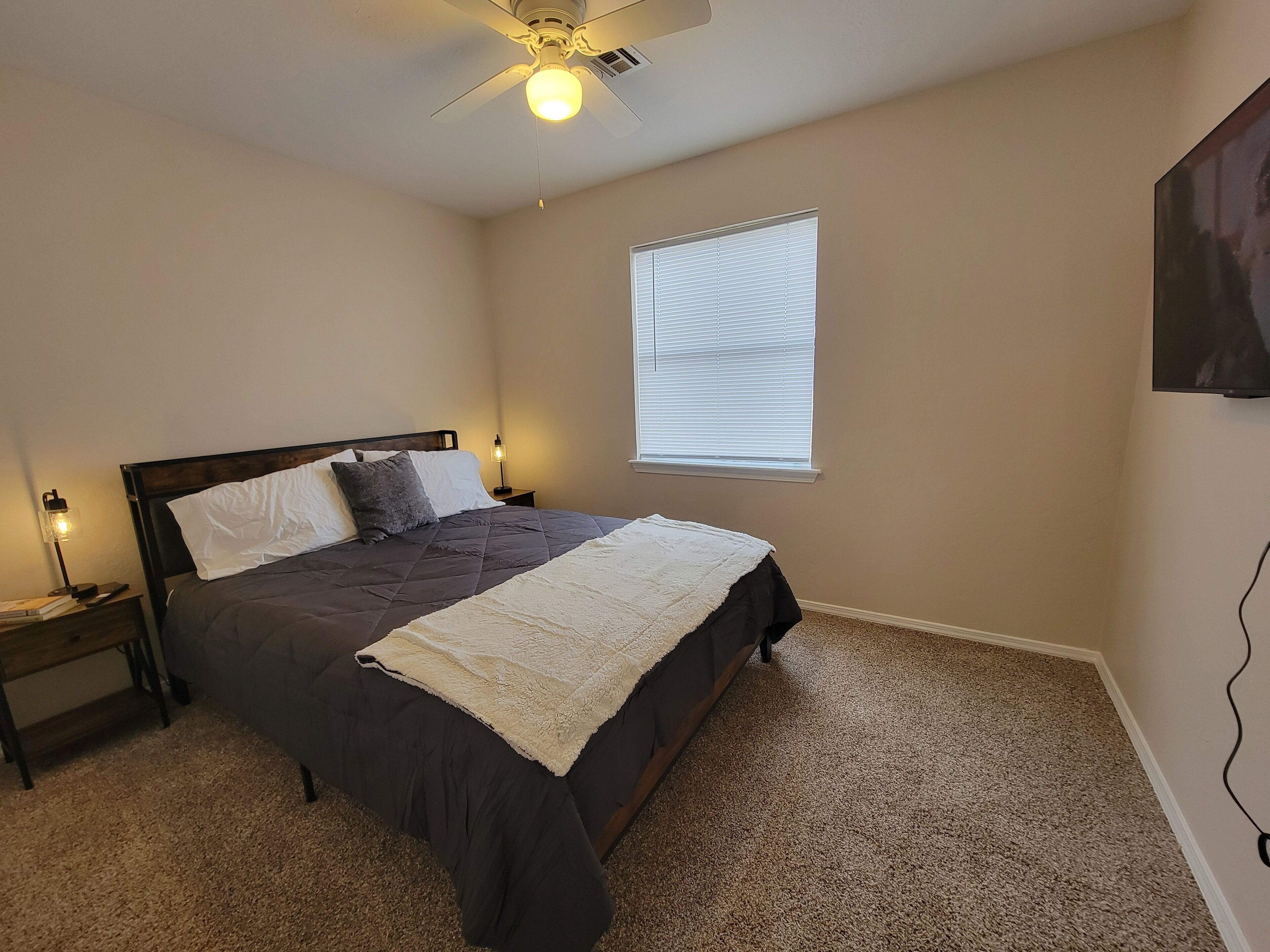 A bedroom with a bed, a ceiling fan, a window with blinds, and a nightstand with a lamp.