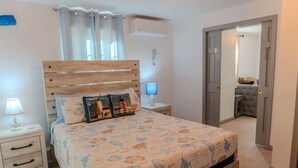 Een slaapkamer met een houten bed, een nachtkastje met een lamp, een wandairconditioner en een deur naar een andere kamer.