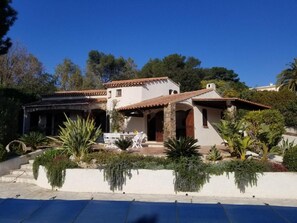Una casa de dos pisos con un patio, mobiliario exterior y un jardín con diversas plantas.