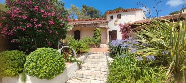 A traditional Mediterranean-style building with a stone pathway leading to a door, surrounded by lush greenery and flowering plants.