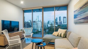 A modern living room with a large TV, comfortable seating, a small table, and a city view through floor-to-ceiling windows.