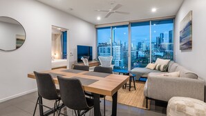 A modern living room with a city view, a dining area, and a bedroom with a cityscape visible through the window.