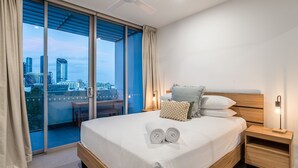 A modern bedroom with a city view, a bed with white linens, a wooden headboard, and a nightstand with a lamp.
