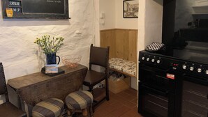 A cozy corner with a wooden table, two plaid cushioned stools, a vase with yellow flowers, a chalkboard menu, and a black oven. - Cottage - Sleeps 4 - Fireplace - Internet (Penzance)