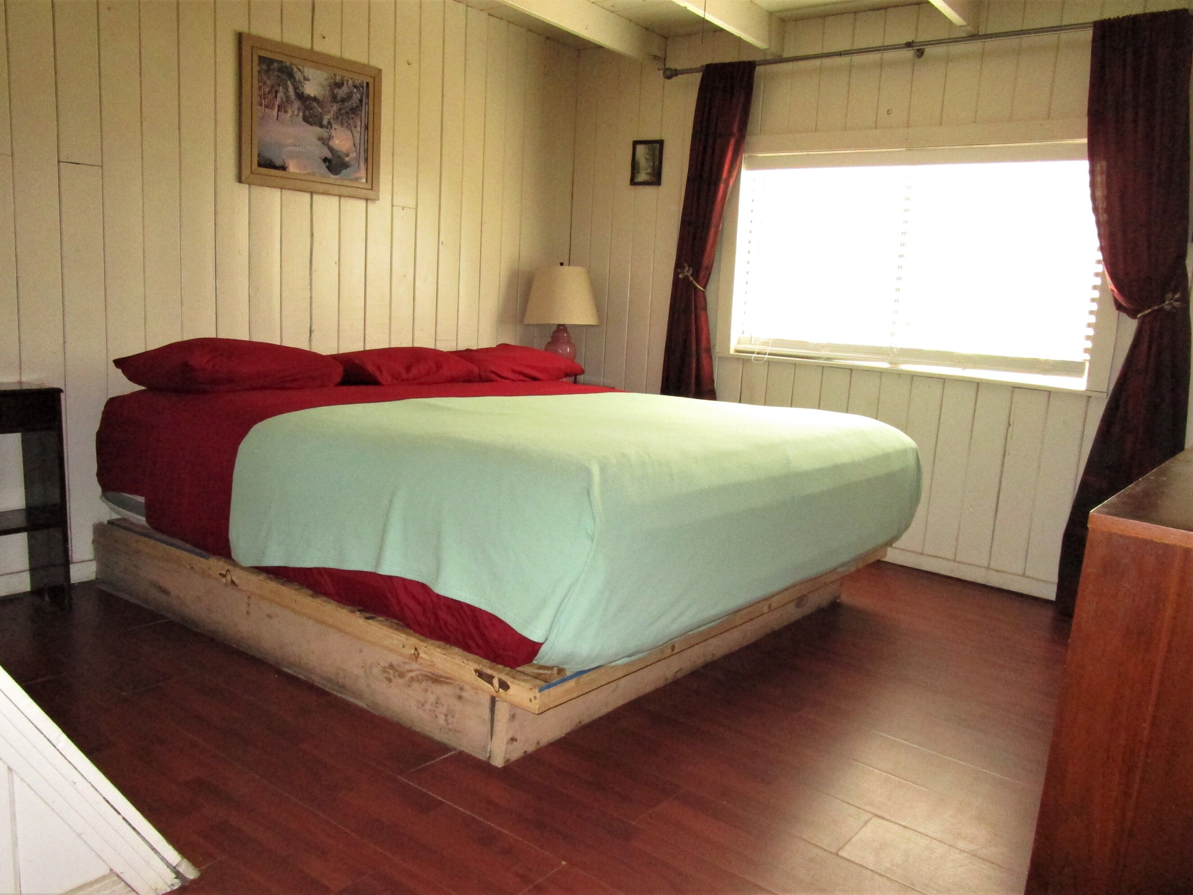 A single-bed room with a wooden frame, a red and green bedspread, a nightstand, a lamp, a window with curtains, and a framed picture on the wall.