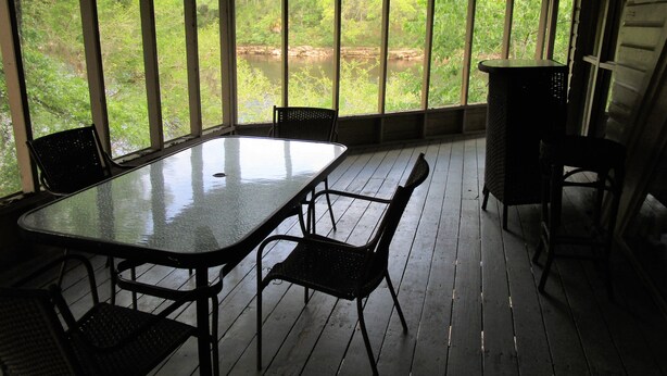 Un espace extérieur abrité, aménagé pour les repas, avec une table et des chaises, de grandes fenêtres et une vue sur la verdure.