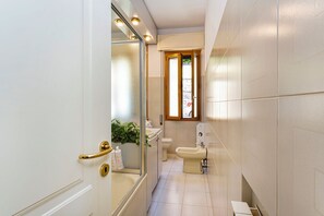 A narrow bathroom with a toilet, bidet, and a small window.