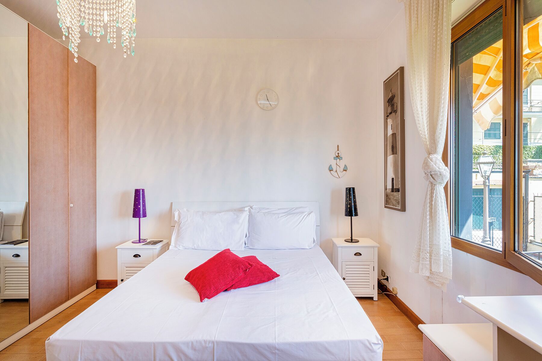 A bedroom with a large bed, two bedside tables, a chandelier, a clock, and a window with curtains.