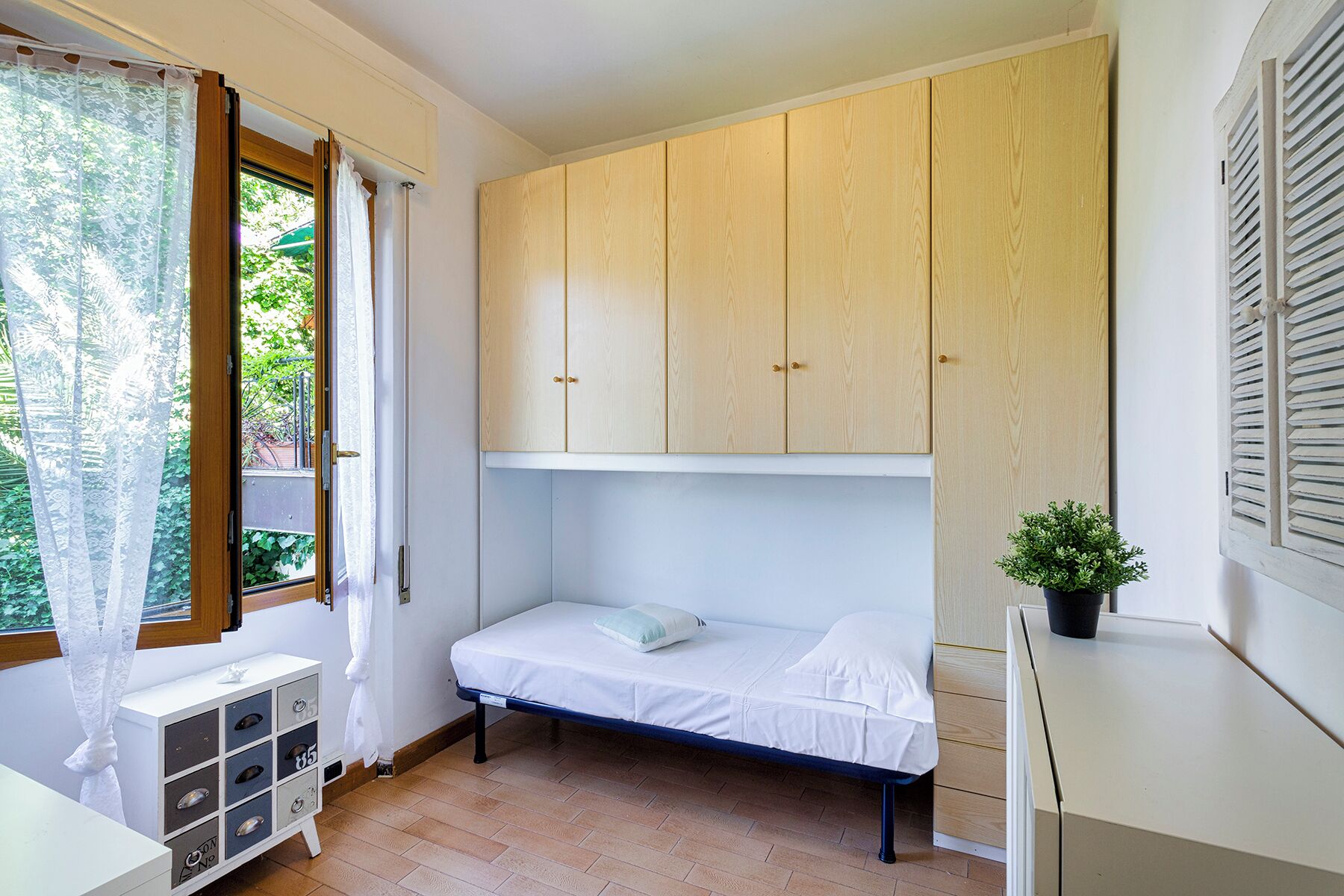 Une petite chambre avec un lit simple, un placard en bois au-dessus du lit, une fenêtre avec des rideaux et une petite plante sur le rebord de la fenêtre.