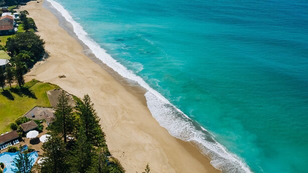 An aerial view of a beach with a clear turquoise sea, sandy shore, and a residential area with houses and a swimming pool. - Ciao Korora - The Ultimate Coffs Coast Getaway (Korora)