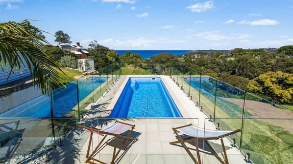 A pool with a clear view of the ocean, surrounded by lush greenery and a house in the background.