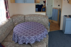 A room with a patterned sofa, a small table with a tablecloth, a kitchenette area, and a door leading to another room. - 3bed-sleeps6-pet Friendly-familyfriendly (Mablethorpe)