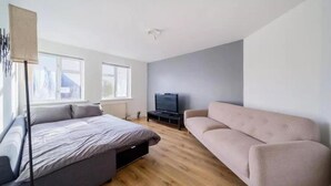 A modern, well-lit room with a bed, a sofa, a TV, and large windows. - 2br-sleeps6-parking-garden-nearlondon (Sunbury on Thames)