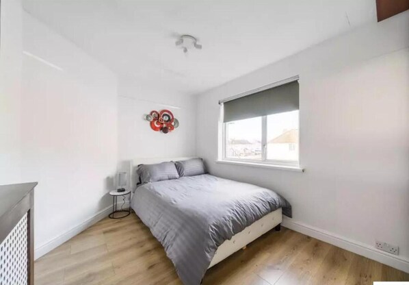 A bedroom with a bed, a small bedside table, a wall decoration, and a window with a view. - 2br-sleeps6-parking-garden-nearlondon (Sunbury on Thames)