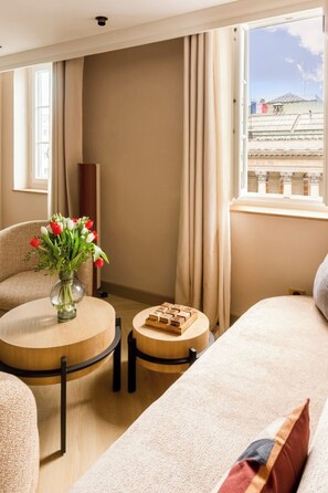 A hotel room with a sofa, a small round table with a vase of flowers, a wooden side table, and a view of buildings through a window. - Merveil - Signature Suite - Louvre - Bourse II (Paris)